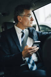 Geschäftsmann mit Smartphone schaut durch das Fenster, während er im Taxi sitzt - MASF18791