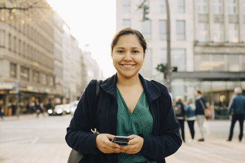 Porträt einer lächelnden Unternehmerin mit Telefon in der Stadt stehend - MASF18805