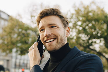 Smiling businessman talking through smart phone while looking away in city - MASF18806