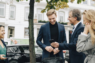 Lächelnder Unternehmer, der einem männlichen Kollegen sein Handy zeigt, während er neben seinen Kollegen in der Stadt steht - MASF18818
