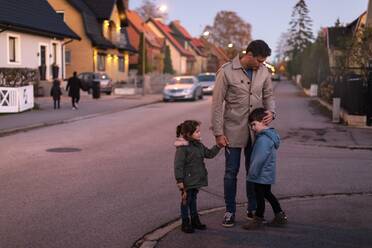 Vater tröstet traurigen Sohn, während er mit Tochter auf dem Fußweg bei Sonnenuntergang die Hände hält - MASF18823