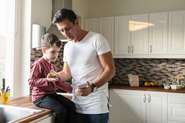 Vater zeigt seinem Sohn das Smartphone mit dem Essen, während er an der Küchentheke steht - MASF18840