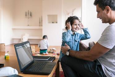 Lächelnde Tochter mit Kopfhörern, die neben ihrem Vater steht, während ihr Bruder im Hintergrund zu Hause spielt - MASF18843