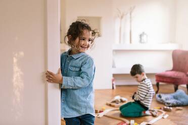 Smiling girl standing by wall while brother playing in background at home - MASF18845