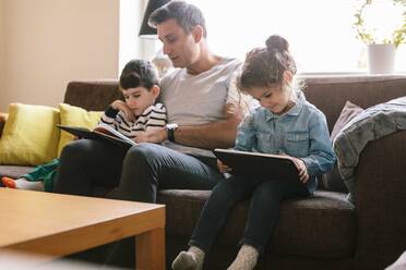 Vater liest seinem Sohn ein Buch vor, während die Tochter auf dem Sofa zu Hause ein digitales Tablet benutzt - MASF18856