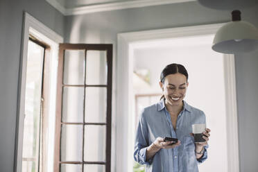 Glücklicher Designprofi mit Getränk und Smartphone im Home Office stehend - MASF18868