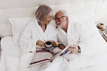 Ältere Frau, die einen Mann ansieht, der eine Zeitung hält, während sie einen Kaffee auf dem Bett im Hotel trinkt - MASF18881