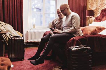 Lächelndes homosexuelles Paar, das auf sein Smartphone schaut, während es auf dem Bett im Hotelzimmer sitzt - MASF18888