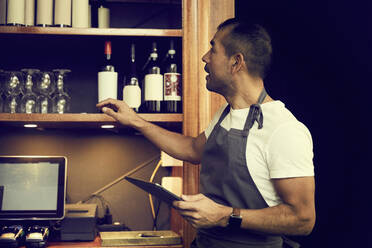 Männlicher Besitzer mit digitalem Tablet, der in einem Café steht und sich Weinflaschen ansieht - MASF18914