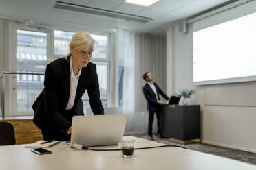 Businesswoman using laptop while colleague standing by flat screen during global conference meeting in office - MASF18943