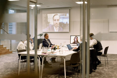 Geschäftsleute diskutieren in einer Sitzung während einer Webkonferenz durch Glas im Büro gesehen - MASF18971