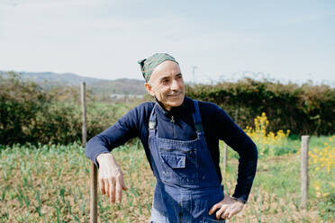 Smiling man wearing dungarees and bandana standing in vegetable garden, leaning on wooden pole. - CUF55613