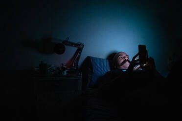 Bald man with moustache lying in bed, using mobile phone while self isolating during Corona crisis. - CUF55629