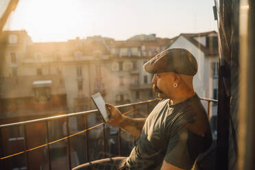 Mann mit Schnurrbart und flacher Kappe sitzt auf einem Balkon und benutzt ein Mobiltelefon, während er sich während der Corona-Krise selbst isoliert. - CUF55631