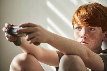 Junge mit roten Haaren sitzt in einem sonnigen Zimmer auf dem Boden und hält den Controller einer Spielkonsole. - CUF55651