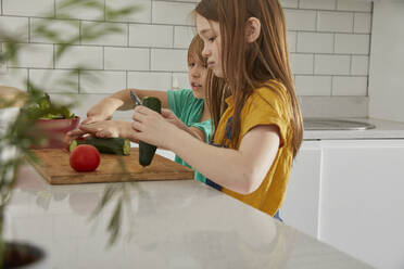 Boy and girl standing in a kitchen, cutting English cucumber and tomato. - CUF55652
