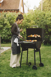 Volle Länge Seitenansicht der älteren Frau bereitet Mahlzeit auf Grill während Abendessen Partei im Hinterhof - MASF18979