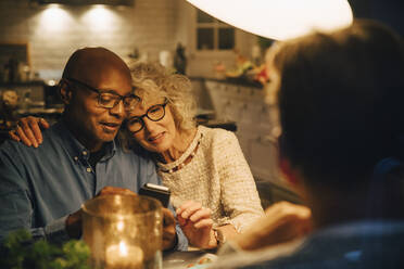 Bald senior sharing smart phone with woman while sitting at illuminated dining table - MASF18987