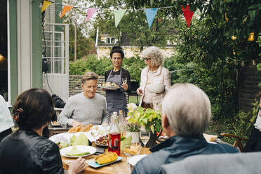 Aktive Seniorinnen bringen Freunden, die während eines Gartenfestes am Esstisch sitzen, das Essen - MASF19003