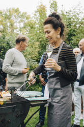Lächelnde ältere Frau mit Getränk in der Hand beim Grillen mit Freunden im Hinterhof - MASF19008