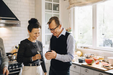 Lächelnder Mann und Frau teilen sich ein Smartphone, während sie in der Küche stehen - MASF19014