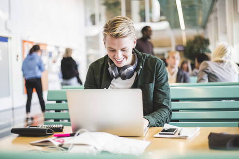 Lächelnder junger männlicher Student mit Laptop in der Cafeteria einer Universität - MASF19120