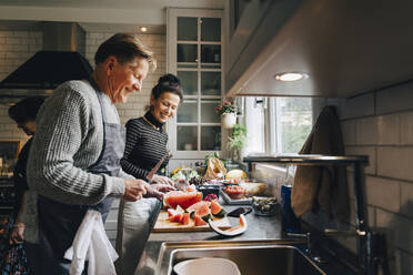 Smiling senior male and female friends cutting fruits at kitchen counter - MASF19156