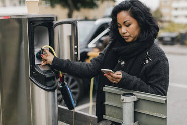 Mid adult woman using smart phone while charging car at station - MASF19163