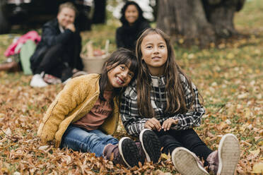 Portrait of smiling sisters sitting on autumn leaves during picnic with parents - MASF19168