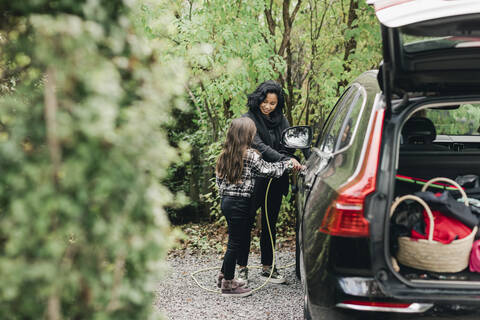 Eine erwachsene Frau bringt ihrer Tochter bei, wie man ein Elektroauto an einer Ladestation auflädt, lizenzfreies Stockfoto