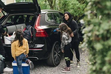 Woman guiding daughter in charging electric car while family loading luggage - MASF19186