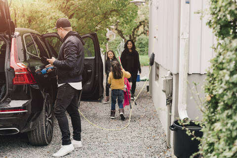 Mann lädt Elektroauto auf, während Familie Gepäck einlädt, lizenzfreies Stockfoto