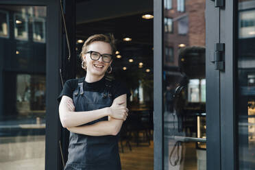 Porträt eines lächelnden androgynen Besitzers, der mit verschränkten Armen am Eingang eines Cafés steht - MASF19208