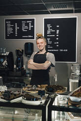 Portrait of smiling barista standing with arms crossed by food variation on counter in illuminated cafeteria - MASF19209