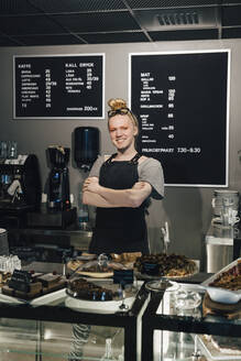 Porträt eines lächelnden Barista, der mit verschränkten Armen neben einer Essensvariation auf dem Tresen einer beleuchteten Cafeteria steht - MASF19209