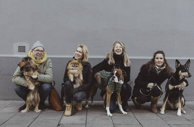 Portrait of smiling pet owners with dogs on footpath against wall in city - MASF19245