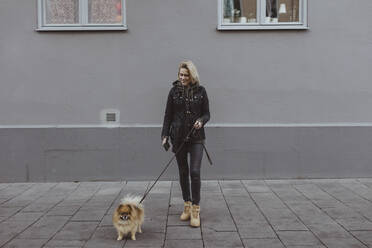 Lächelnde Frau in voller Länge mit pommerschem Hund auf einem Fußweg vor einem Gebäude stehend - MASF19247