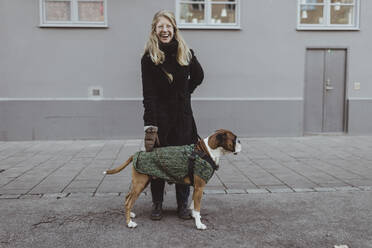 Fröhliche junge Frau steht mit Boxer vor einem Gebäude in der Stadt - MASF19255