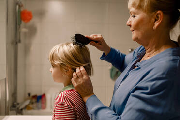 Side view senior woman combing hair of grandson in bathroom at home - MASF19327