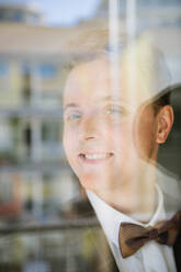 Handsome young male in suit and bow tie smiling and looking at camera while standing behind window glass - ADSF00162