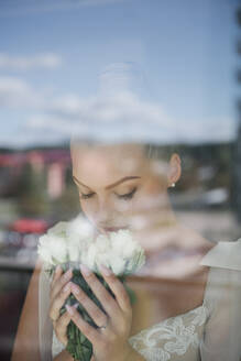 Pretty bride in white gown standing at window and smelling bunch of flowers - ADSF00164