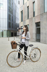 Businesswoman drinking water while standing with bicycle on city street - VEGF02459