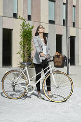 Relaxed businesswoman standing with bicycle on city street during sunny day - VEGF02461