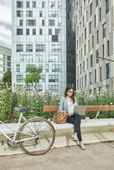 Female commuter using laptop while sitting on bench in city - VEGF02462