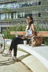 Cheerful businesswoman sitting with laptop on bench in city during sunny day - VEGF02464