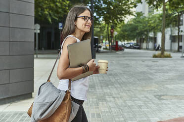 Nachdenkliche Geschäftsfrau mit Laptop und Einwegglas beim Spaziergang auf der Straße - VEGF02468