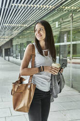 Nachdenkliche Geschäftsfrau, die ein Einwegglas in der Hand hält, während sie in einem Durchgang vor einem Bürogebäude steht - VEGF02471
