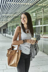 Thoughtful businesswoman holding disposable glass while standing at passage outside office building - VEGF02471