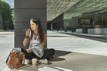 Geschäftsfrau, die ein Smartphone benutzt, während sie auf der Kolonnade vor einem Bürogebäude sitzt - VEGF02473