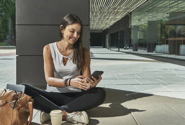 Geschäftsfrau, die ein Smartphone benutzt, während sie an einem sonnigen Tag auf der Kolonnade sitzt - VEGF02474